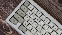 Load image into Gallery viewer, GMK MTNU Beige keycaps on white 65% keyboard, angled view from front left on dark wood surface